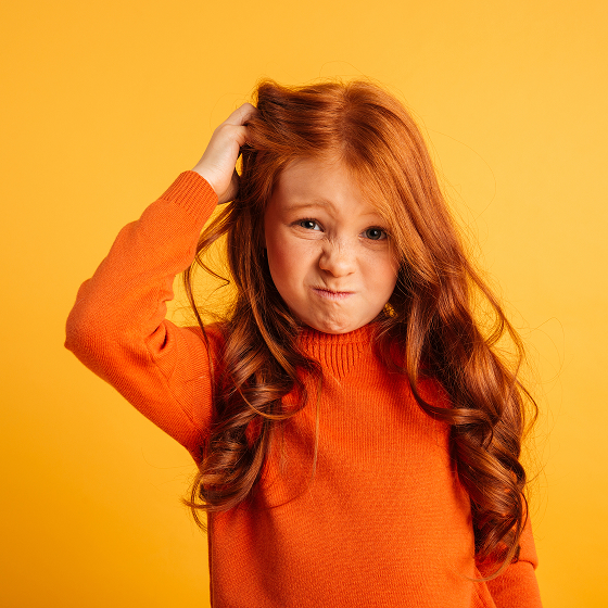 A little girl about age 8, white with red hair and wearing an orange sweater, scratches her head in confusion