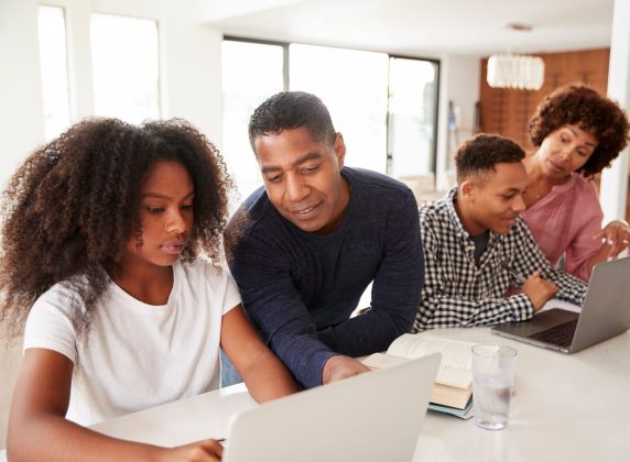 Two Black parents, a man and woman, helping their two teenage children on their computers, a boy and a girl