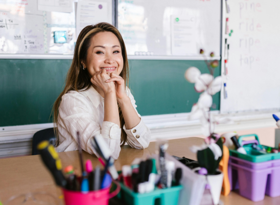 A female Asian teacher sitting at a desk smiling into the camera