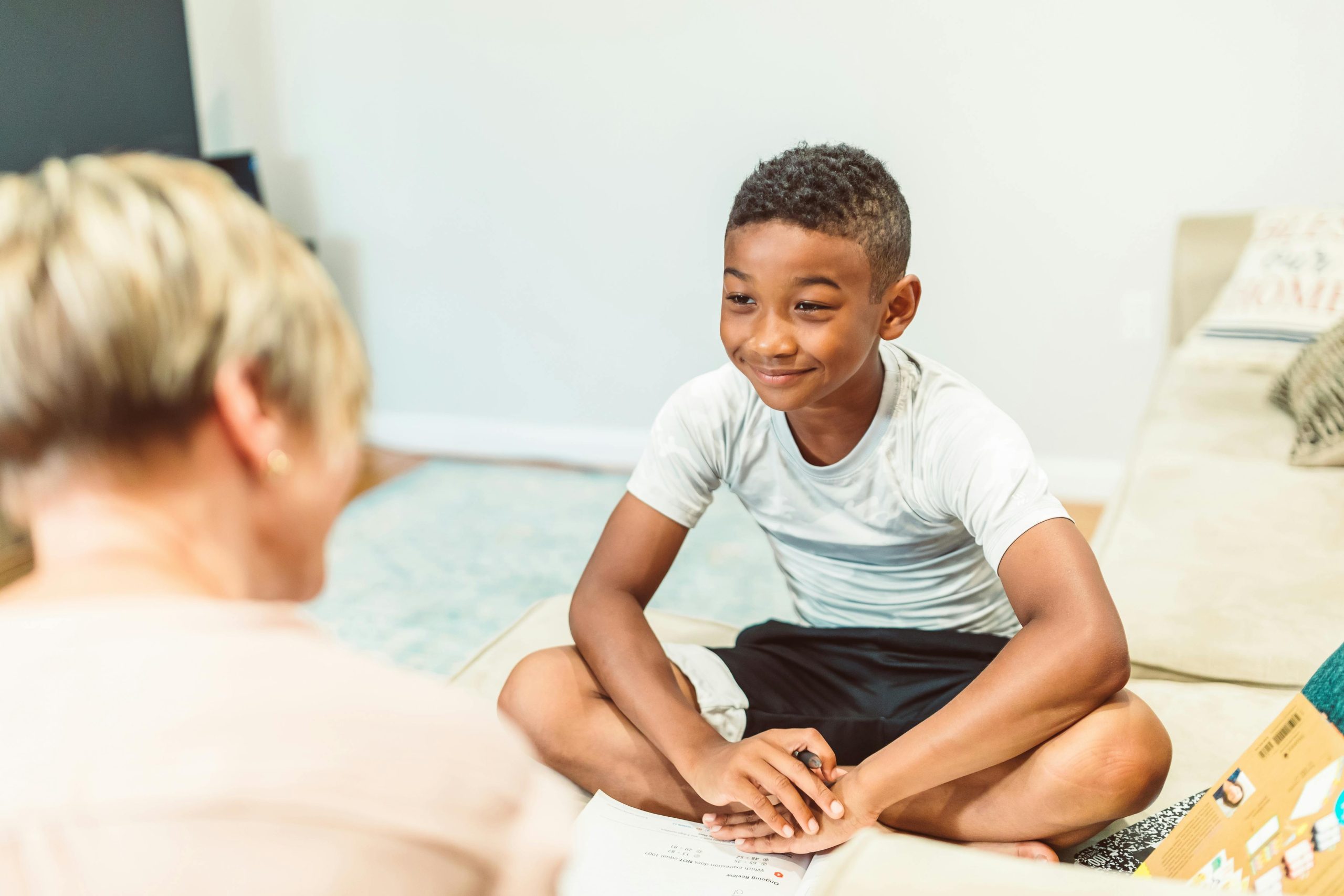 A young Black boy sits on the floor at school, smiling while learning and interacting with his teacher and peers