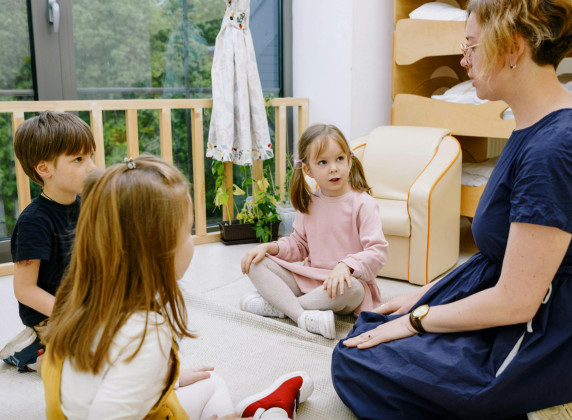 An early learning teacher, a white woman with blonde hair, sitting in a circle with three other white children