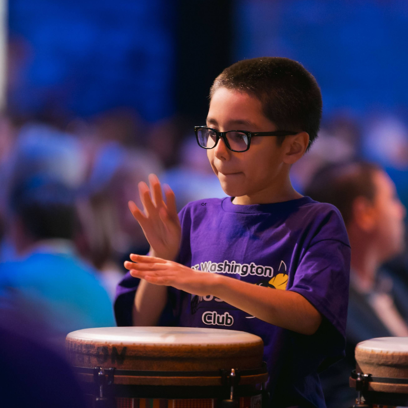 A little elementary school boy plays the drums at a Frameworks event wearing a purple school T-shirt and glasses