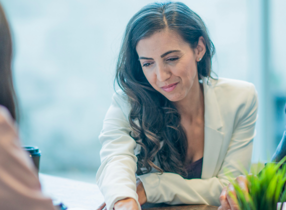 White woman with long brown hair sitting at a desk talking to another leader you can't see