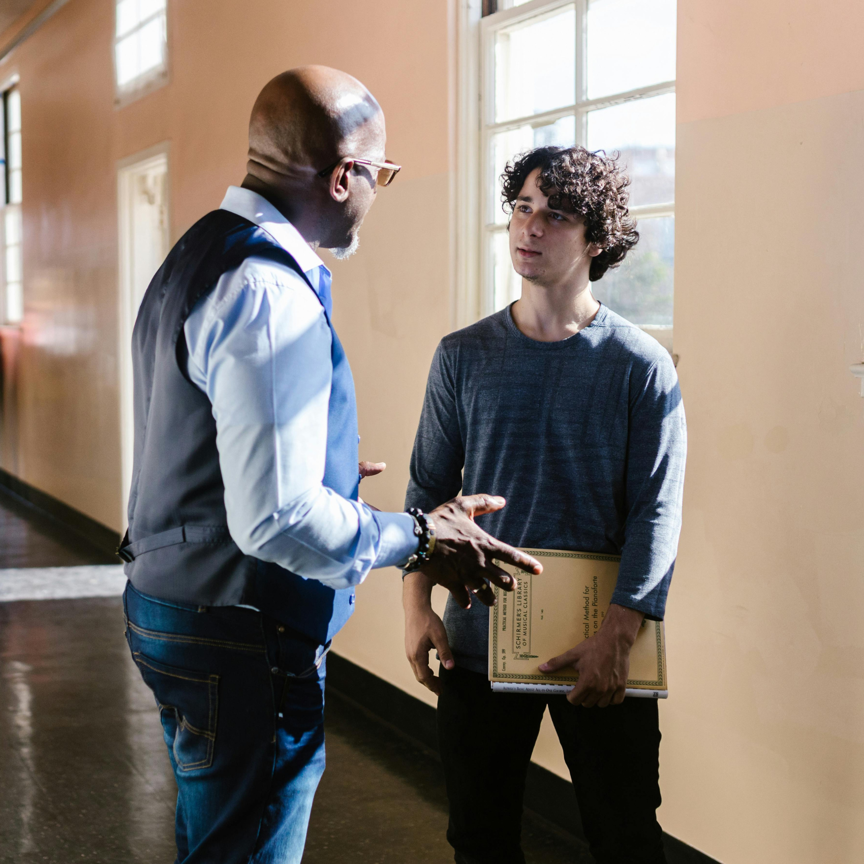 A Black male teacher, bald wearing a blue button down shirt and vest, talks with a white teen boy with brown curly hair wearing a grey-blue sweater in the hallway at school